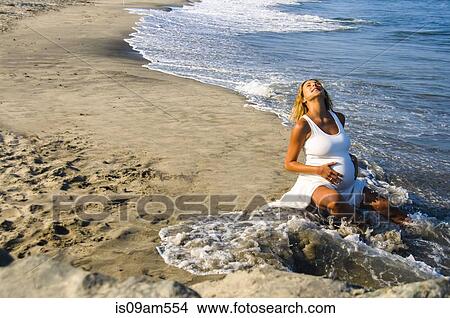 Pregnant mature woman sitting in ocean waves on beach whilst touching stomach View Large Photo Image Picture - Pregnant mature woman sitting in ocean waves on beach whilst touching stomach. Fotosearch