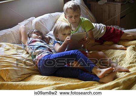 Three young children relaxing on bed using digital tablet View Large Photo Image Stock Image - Three young children relaxing on bed using digital tablet. Fotosearch