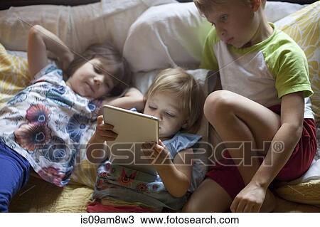 Stock Image - Three young children relaxing on bed using digital tablet. Fotosearch