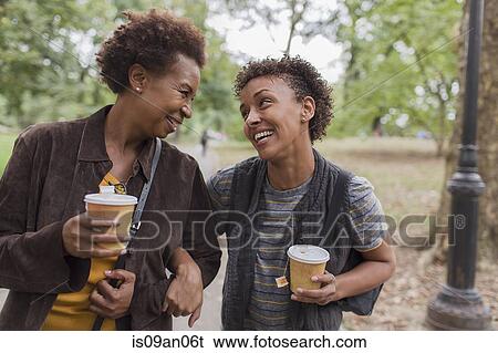 Stock Photograph - Two mature female friends drinking takeaway coffee whilst strolling in park. Fotosearch