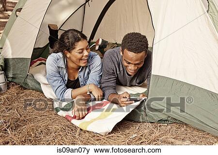 Stock Photo - Young couple lying in tent entrance reading texts on smartphone. Fotosearch