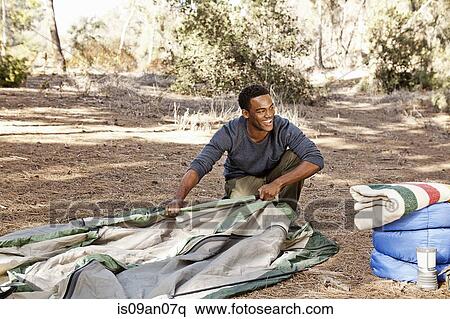 Stock Photography - Young man kneeling in forest putting up tent. Fotosearch