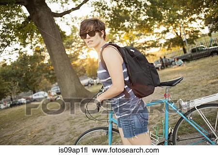 Stock Image - Female cyclist pushing bicycle through park. Fotosearch