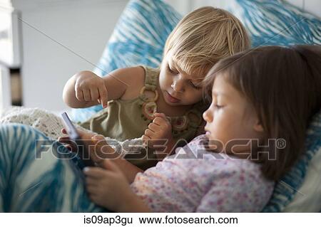 Stock Photograph - Girl and toddler sister sitting up in bed using digital tablet. Fotosearch