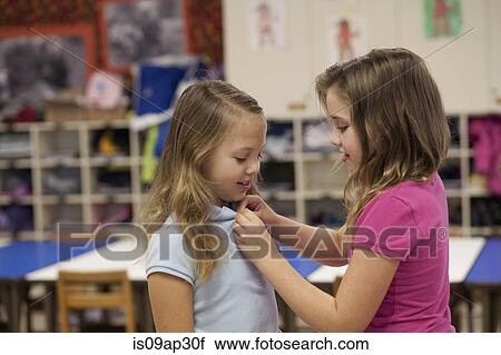 Stock Image - Girl buttoning friend's tee shirt in classroom. Fotosearch
