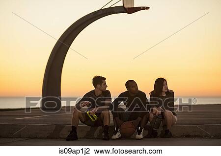 Stock Image - "Group of friends sitting on edge of basketball court, outdoors". Fotosearch