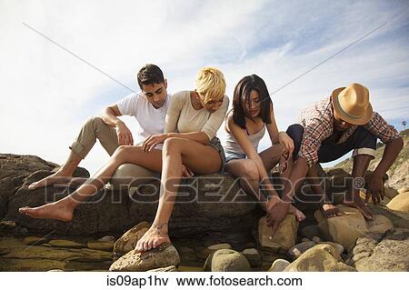 Stock Photo - "Group of friends sitting together on rocks on beach, looking in rock pools". Fotosearch