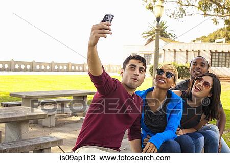 Stock Image - "Group of friends sitting together, taking self portrait with smartphone". Fotosearch