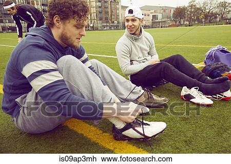 Stock Image - Male adult friends tying boot laces on soccer pitch. Fotosearch