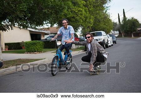 Stock Photo - Male skateboarder skitching on friends moped along suburban road. Fotosearch