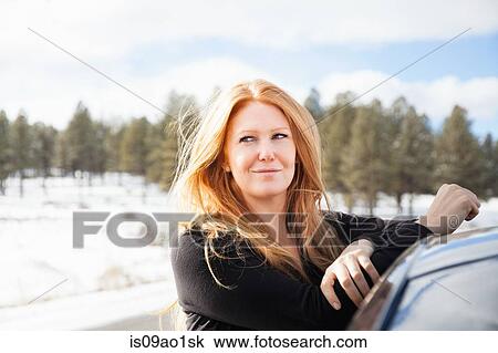 Stock Image - Mid adult woman leaning against car roof on snowy roadside. Fotosearch