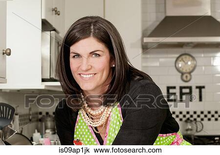 Stock Image - "Mid adult woman wearing apron in kitchen, portrait". Fotosearch