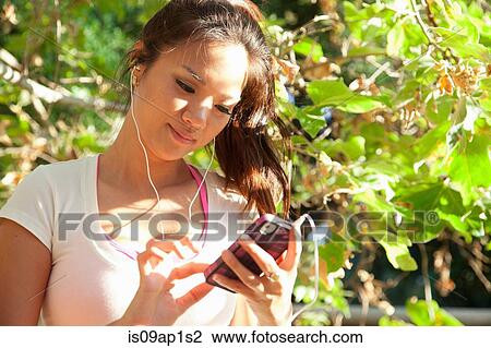 Stock Image - "MId adult woman wearing earphones and holding smartphone, outdoors". Fotosearch