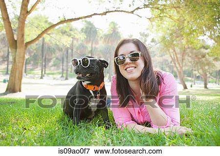 Stock Photo - Portrait of young woman and dog lying in park wearing sunglasses. Fotosearch