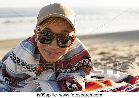Stock Image - "Portrait of young woman sitting on beach, wearing cap and sunglasses". Fotosearch