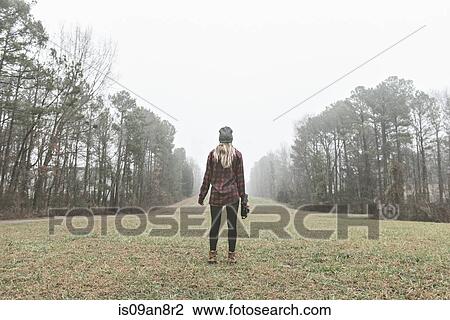 Stock Image - Rear view of young female photographer standing in misty field. Fotosearch