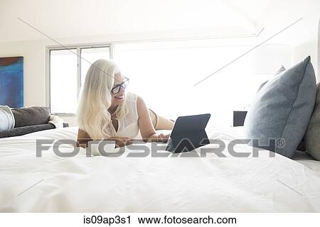 Stock Image - "Senior woman lying on bed, using tablet computer". Fotosearch