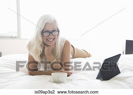 Stock Image - "Senior woman lying on bed, with tablet computer and cup of tea". Fotosearch