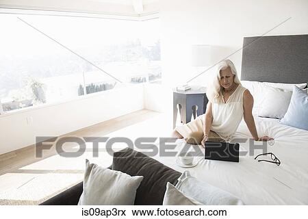 Stock Photo - Senior woman sitting on bed and looking at tablet computer. Fotosearch