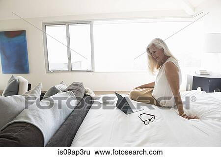Senior woman sitting on bed and looking at tablet computer View Large Photo Image Stock Photograph - Senior woman sitting on bed and looking at tablet computer. Fotosearch