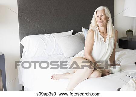 Stock Image - Senior woman sitting on bed with cup of tea. Fotosearch