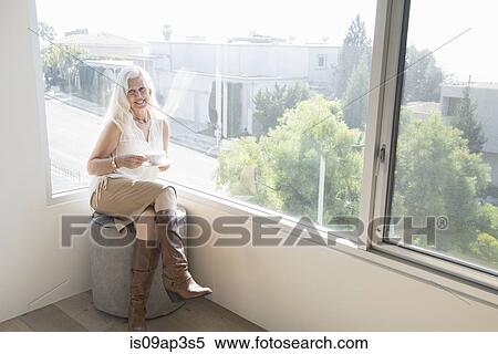 Stock Photography - Senior woman sitting on stool with cup of tea. Fotosearch