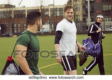 Picture - Three male soccer players carrying gym bags on soccer pitch. Fotosearch