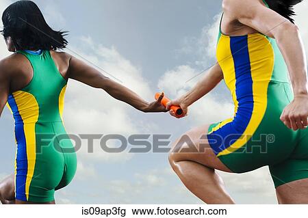 Stock Image - Two female relay athletes exchanging baton whilst running. Fotosearch