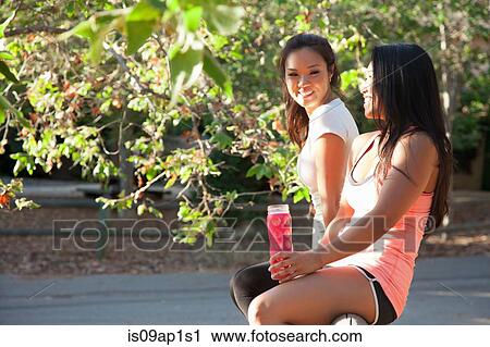Stock Image - "Two mid adult women sitting on fence, smiling". Fotosearch