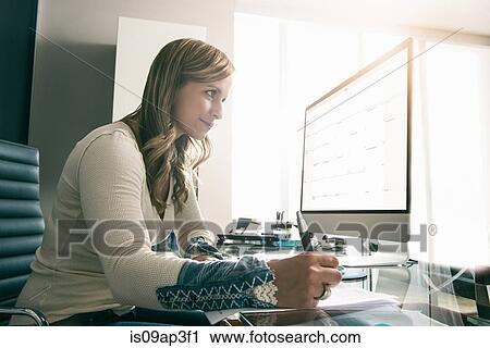 Stock Image - Young businesswoman using reading computer screen whilst making notes. Fotosearch