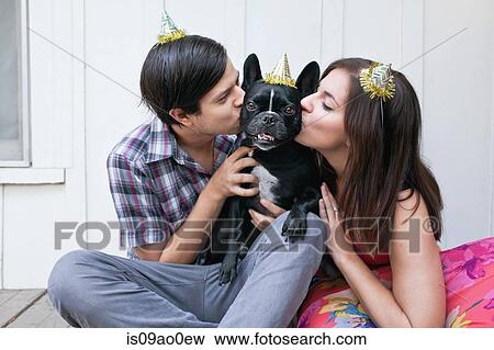 Stock Photo - Young couple kissing dog on cheek and wearing party hats. Fotosearch
