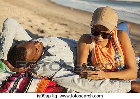 Stock Image - "Young couple lying together on beach, woman looking at smartphone". Fotosearch