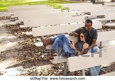 "Young couple sitting together on bench, sharing earphones" View Large Photo Image Stock Image - "Young couple sitting together on bench, sharing earphones". Fotosearch