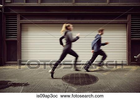 Stock Image - Young man and woman running along city sidewalk. Fotosearch