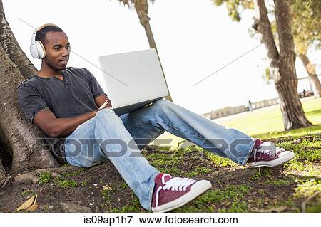 "Young man leaning against tree, wearing headphones and using laptop" View Large Photo Image Stock Photo - "Young man leaning against tree, wearing headphones and using laptop". Fotosearch