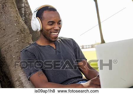 Stock Photo - "Young man leaning against tree, wearing headphones and using laptop". Fotosearch