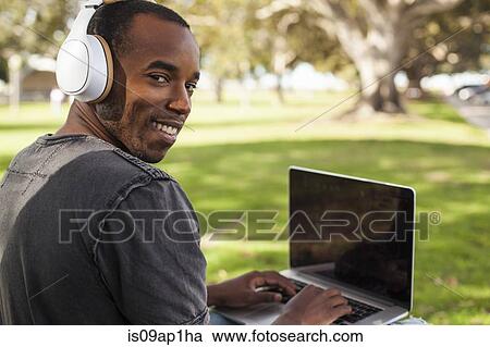 Stock Image - "Young man, outdoors, wearing headphones and using laptop, looking over shoulder". Fotosearch
