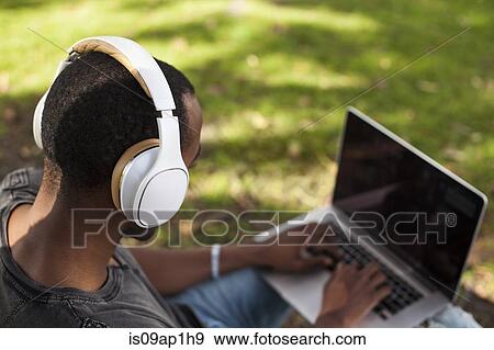 Stock Photo - "Young man, outdoors, wearing headphones and using laptop, rear view". Fotosearch