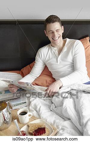 Young man sitting up in bed and laughing whilst reading newspaper View Large Photo Image Stock Photography - Young man sitting up in bed and laughing whilst reading newspaper. Fotosearch