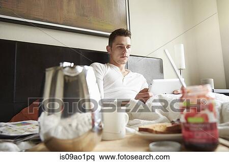 Young man sitting up in bed reading digital tablet View Large Photo Image Stock Photography - Young man sitting up in bed reading digital tablet. Fotosearch