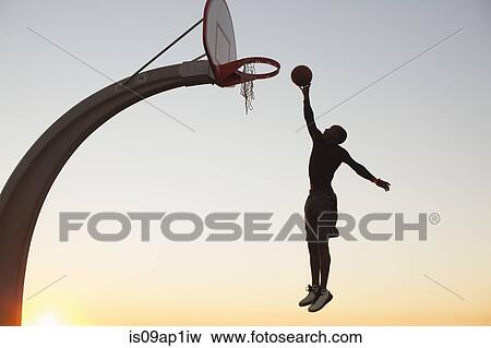 Stock Photo - "Young man with basketball, jumping towards net, outdoors". Fotosearch