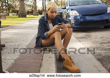 Picture - "Young woman sitting on kerb, using smartphone". Fotosearch