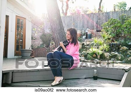 Stock Image - Young woman sitting on patio step petting dog. Fotosearch