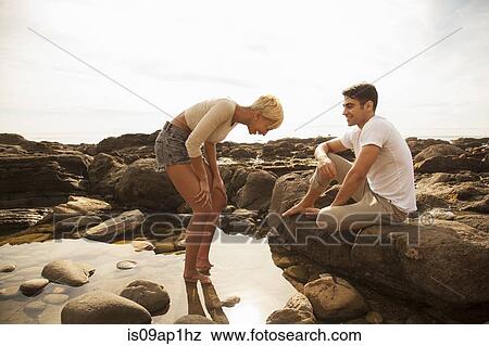 Stock Photo - "Young woman standing in rock pool, man sitting on rocks observing". Fotosearch