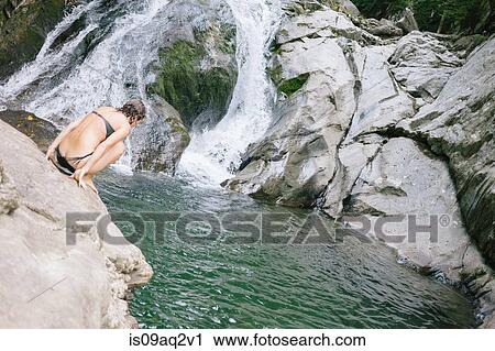 Stock Image - A woman contemplating jumping into a swimming hole in the woods with a waterfall. Fotosearch
