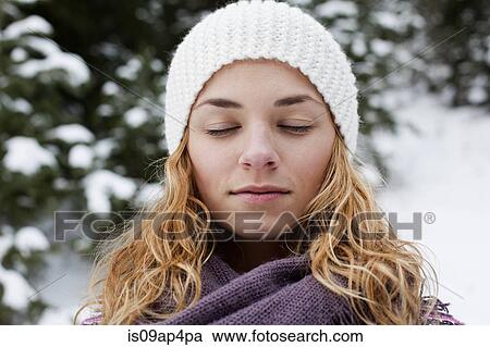 Stock Image - Close up portrait of young woman with eyes closed in front of snow covered fir trees. Fotosearch
