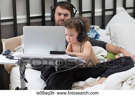 Stock Image - Father watching toddler daughter typing on laptop in bed. Fotosearch