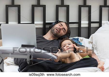 Stock Image - Female toddler listening to headphone music in bed whilst father sleeps. Fotosearch