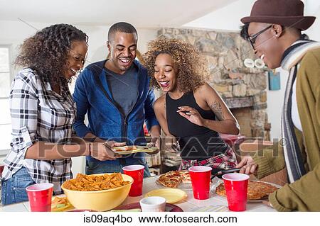 Picture - Four adult friends eating party food in kitchen. Fotosearch