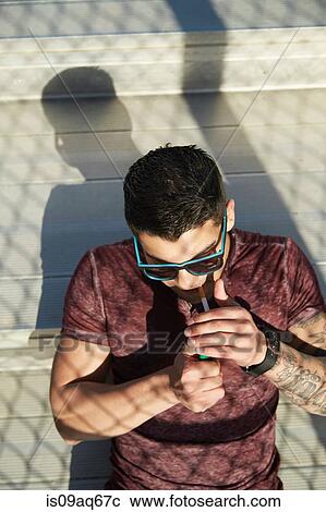 Stock Image - High angle view of young man lighting cigarette on park bench. Fotosearch